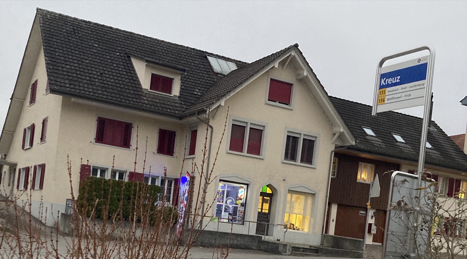 Das Haus an der Hauptstrasse 41 mit dem beleuchteten Schaufenster, vorne rechts das Haltestellenschild Kreuz